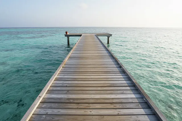 Weathered wooden jetty extends over clear turquoise shallow ocean water at soft sunrise