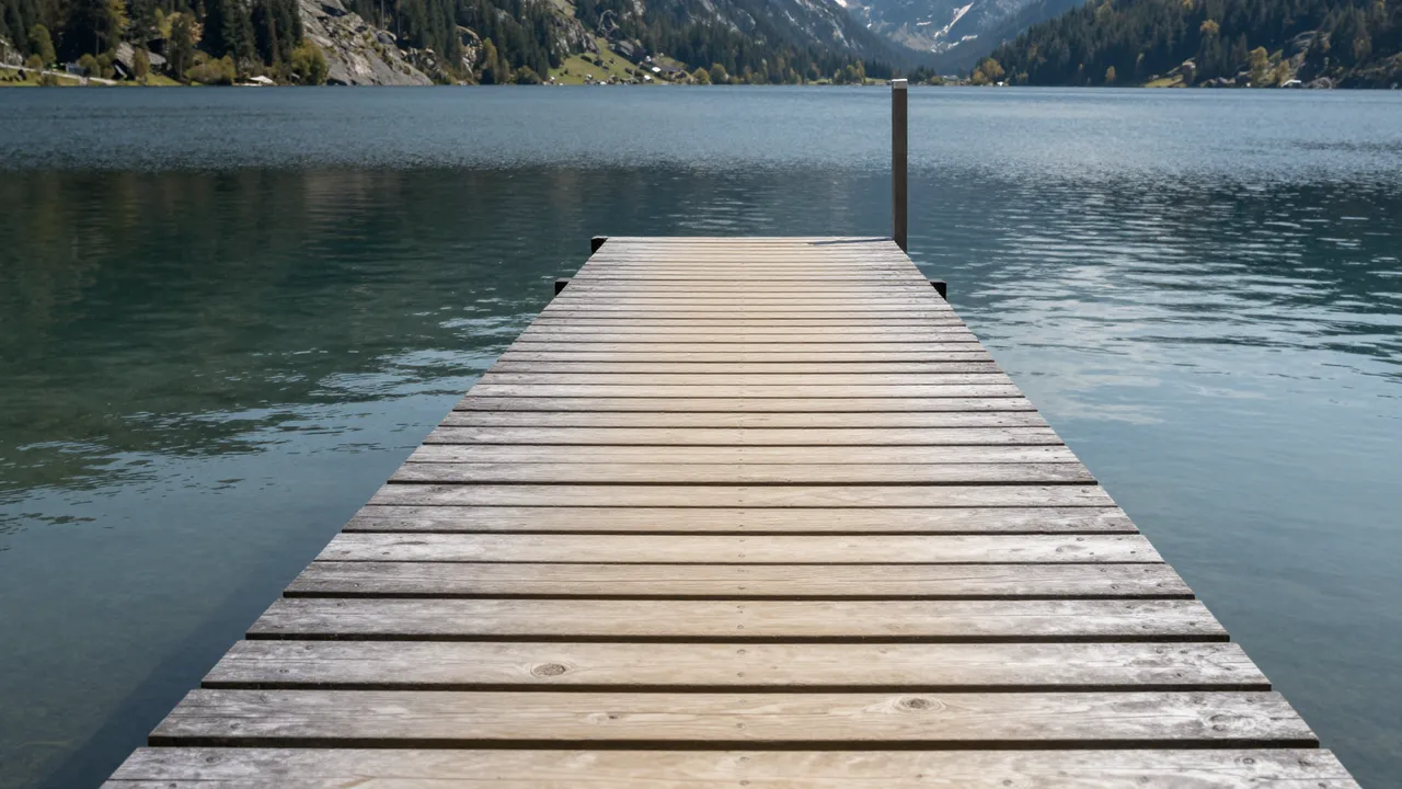 Weathered wooden dock extends out over calm clear lake water toward forested mountain ranges