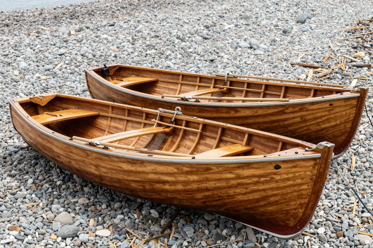 Two polished wooden rowboats rest side by side on a textured pebble shoreline near calm water.