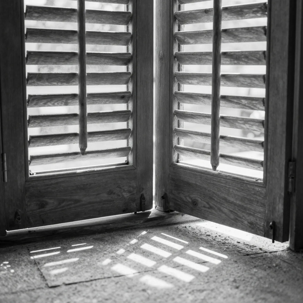 Black and white view of wooden louvered shutters casting angular light patterns onto a textured stone floor
