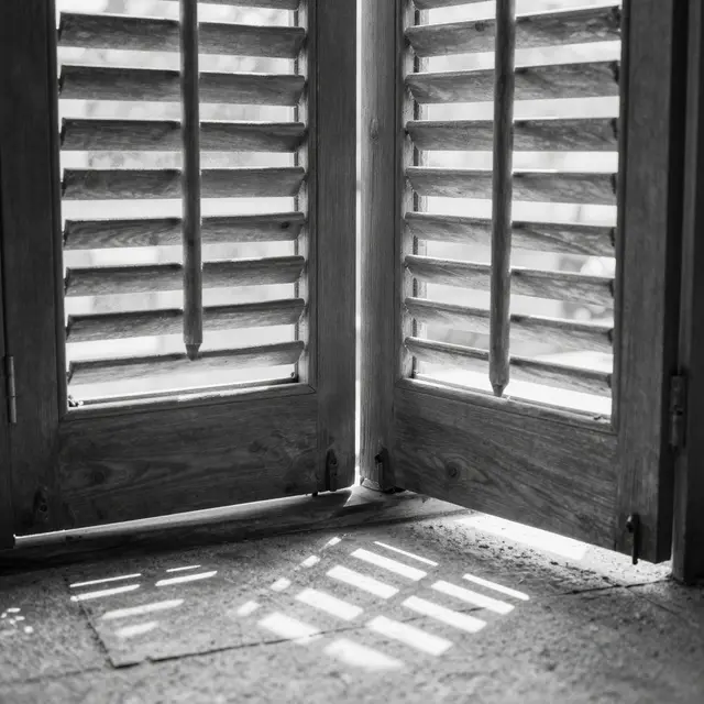 Black and white view of wooden louvered shutters casting angular light patterns onto a textured stone floor