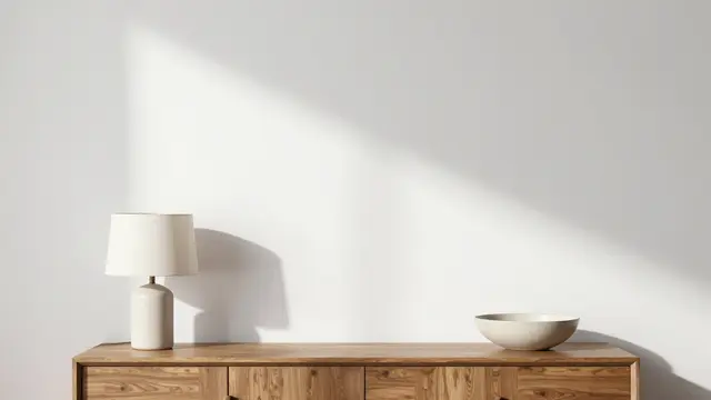 Empty white wall above a wooden sideboard holding a table lamp and ceramic bowl with soft diagonal sunlight.