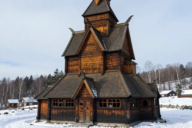 An ornate historic wooden stave church stands in a snow-covered winter landscape under pale overcast skies