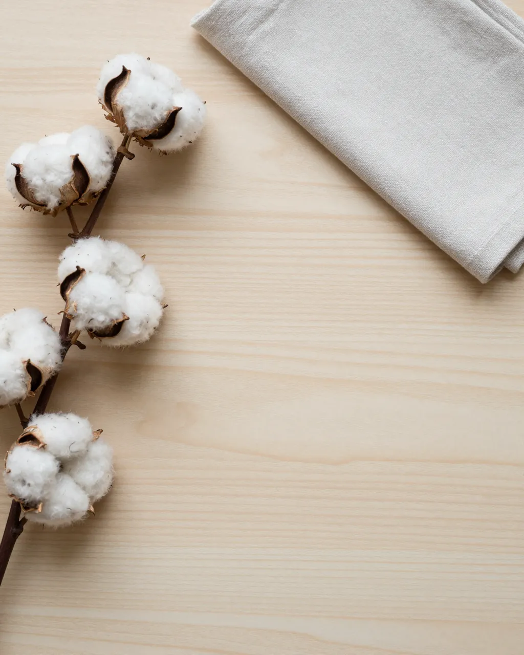 Top view of a pale wooden table with a cotton sprig and folded linen napkin in the upper corners, with empty copy space in