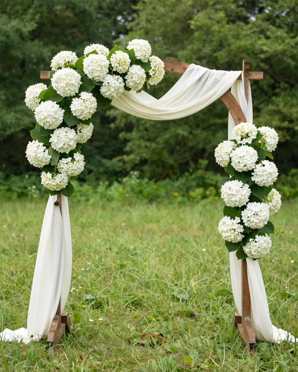 Round wooden wedding arch draped in ivory fabric and decorated with white hydrangeas stands in a sunny grassy meadow