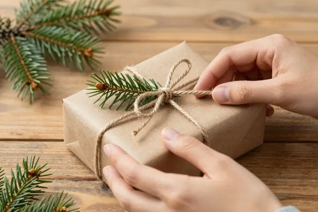 Two hands tie twine around a kraft paper gift box decorated with pine sprigs on a wooden table