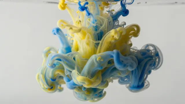 Swirling yellow and blue ink disperses and twists within clear still water against a pale neutral background