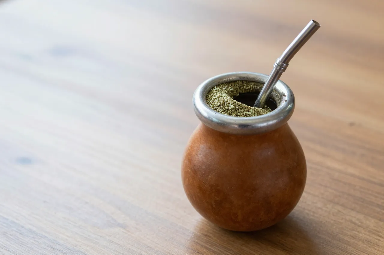 A yerba mate gourd with a metal bombilla straw sits on a polished wooden table with empty copy space to the left.