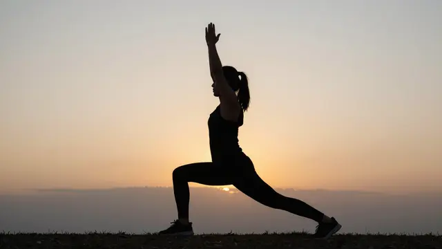 Silhouette of a person holding a lunge yoga pose with raised arms on a hilltop at sunrise