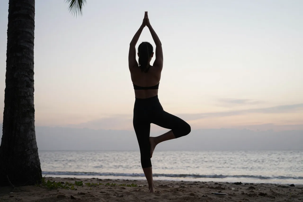 Silhouette of a woman holding yoga tree pose on a sandy beach at soft pale dawn