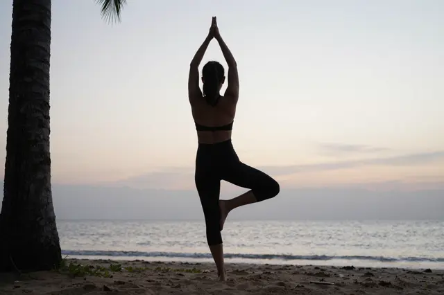 Silhouette of a woman holding yoga tree pose on a sandy beach at soft pale dawn