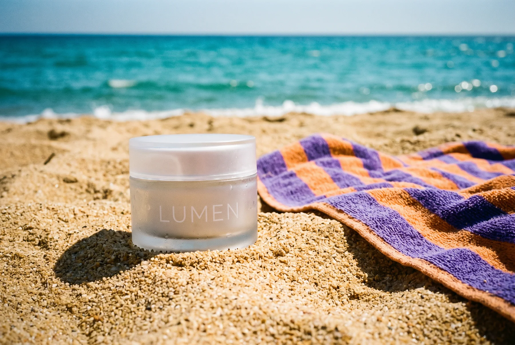 The same LUMEN jar standing in warm sand at noon next to a violet-and-orange striped beach towel, with turquoise sea behind it. An edit made with Nano Banana 2.1 from the product shot above.