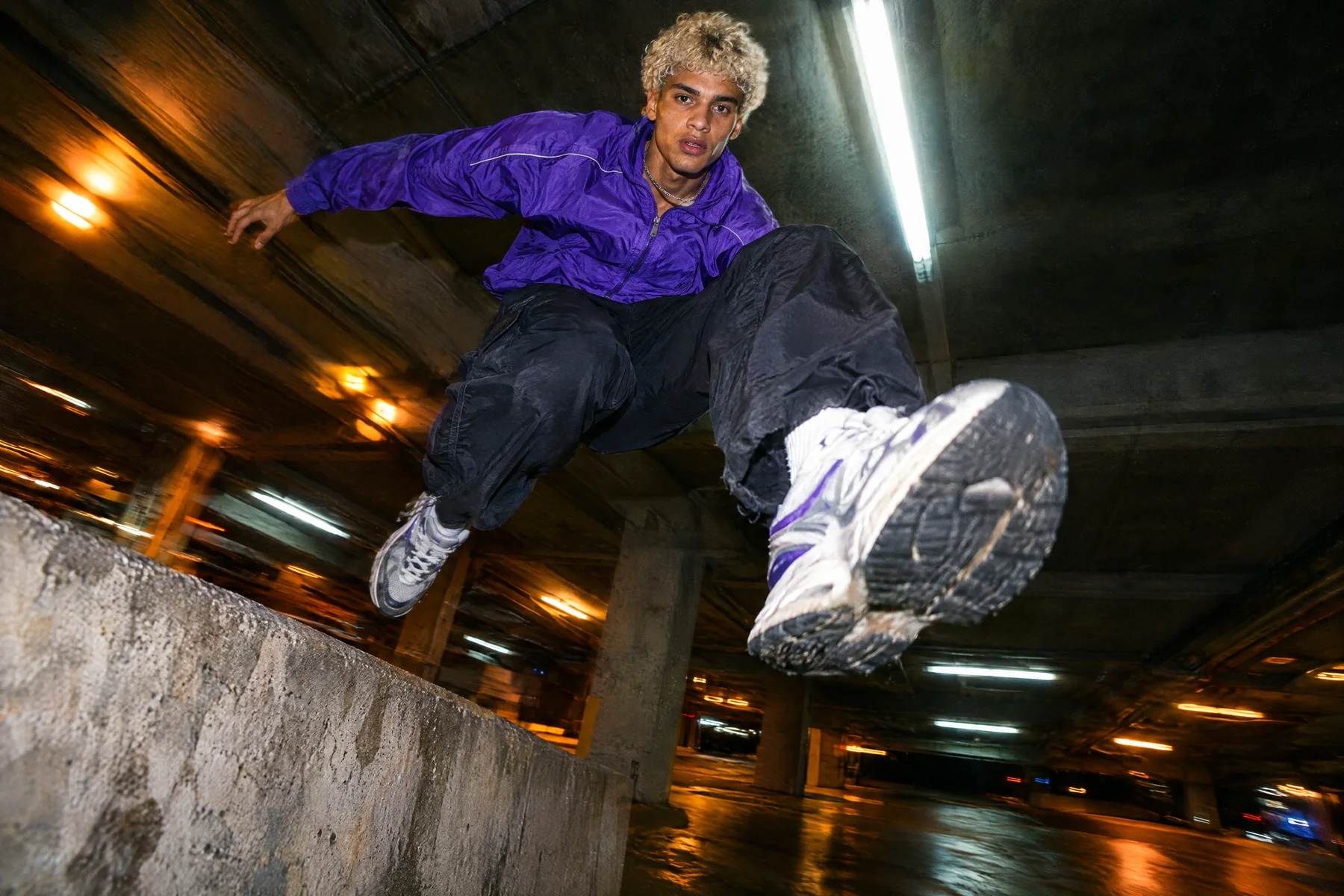A young man with bleached curly hair in an electric-violet track jacket jumps off a concrete ledge in a parking garage at night, one sneaker toward the camera, lit by flash, with fluorescent lights streaking behind him. Made with GPT Image 2.5 Flare.