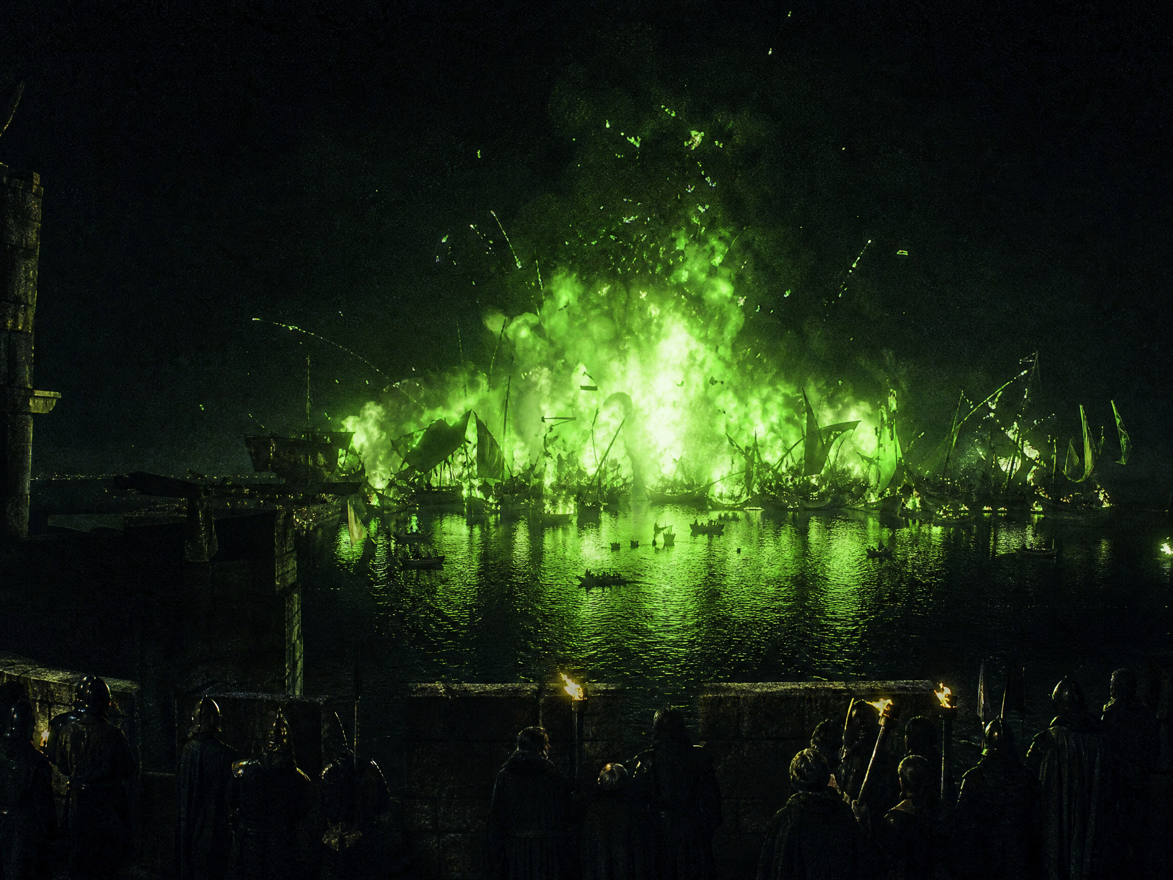 Silhouetted castle defenders watch bright green wildfire explode across a burning enemy fleet on Blackwater Bay at night.