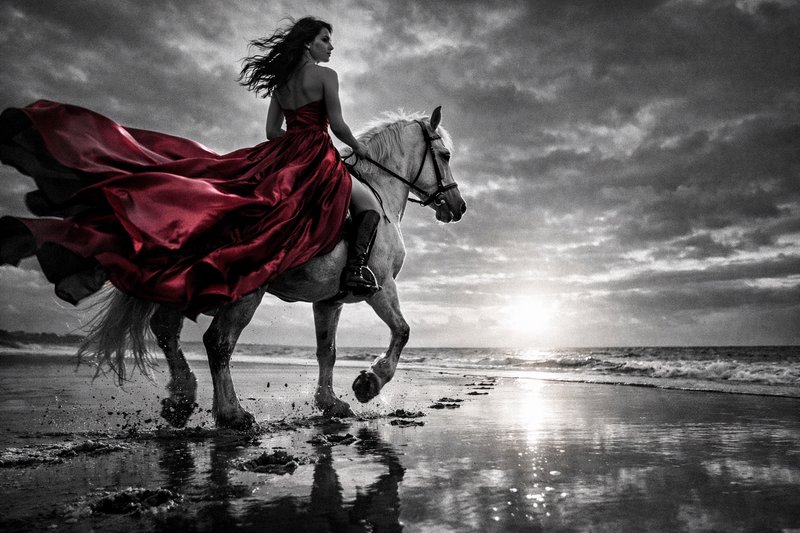 Woman in a flowing red dress rides a white horse along a beach at sunset.