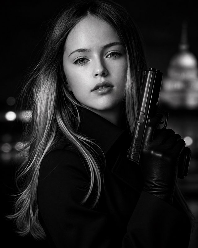 Black-and-white portrait of a young woman in a coat and gloves, holding a pistol in a moody urban setting.