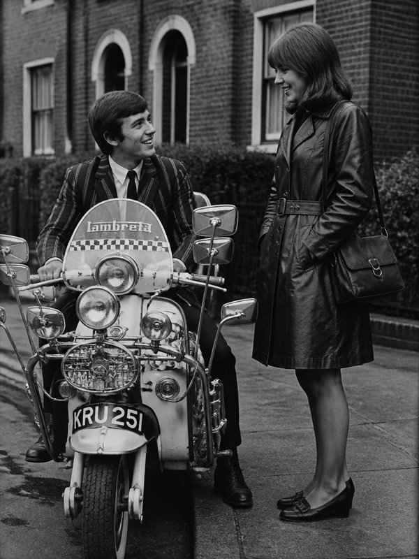 Smiling man in a suit on a vintage Lambretta scooter, talking with a woman in a leather coat beside a brick building.