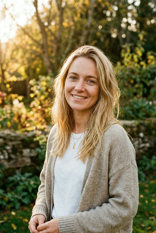 Smiling woman with long blonde hair in a beige cardigan outdoors during golden hour.