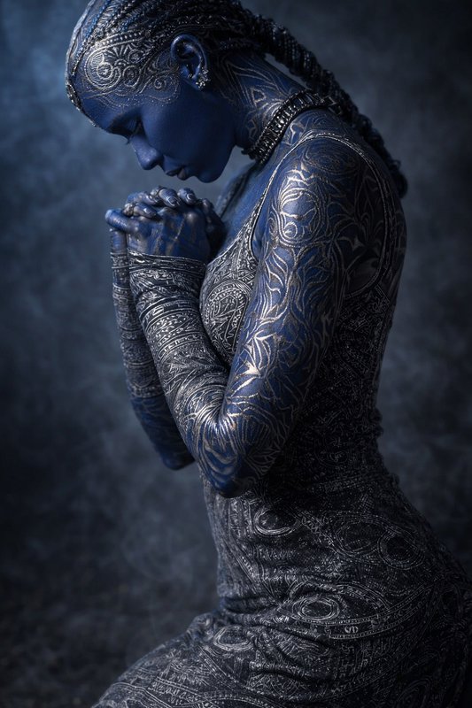Blue body-painted woman with intricate white patterns in a prayer pose.