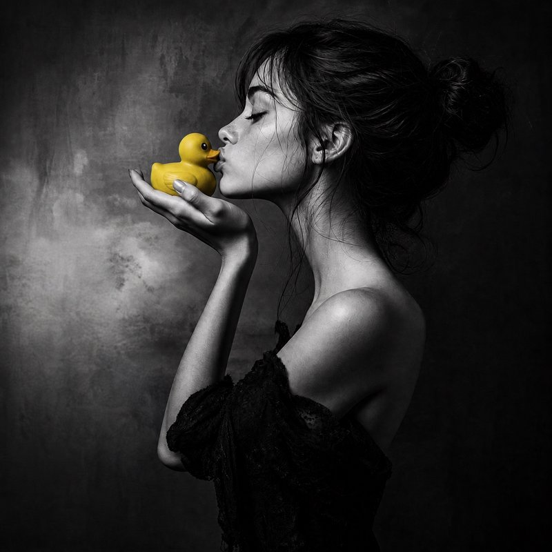 [Side profile artistic portrait of a woman gently kissing a bright yellow rubber duck, selective color monochrome shot]