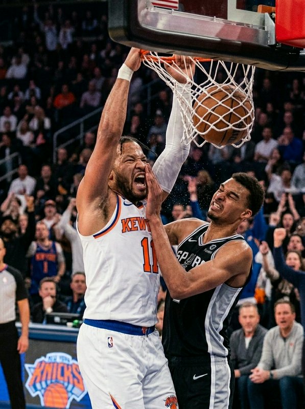Knicks player dunks over Spurs defender at the basket in a crowded arena.