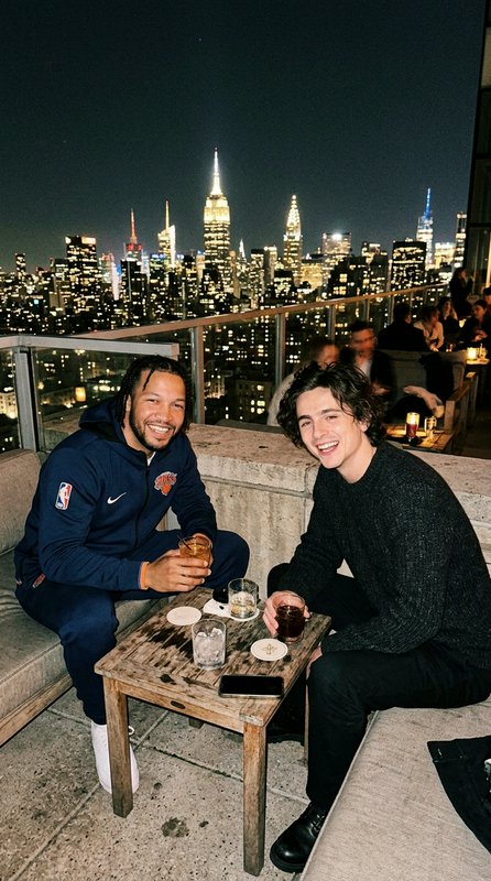 Jalen Brunson and Timothee Chalamet laugh over cocktails at a NYC rooftop bar, with the illuminat...
