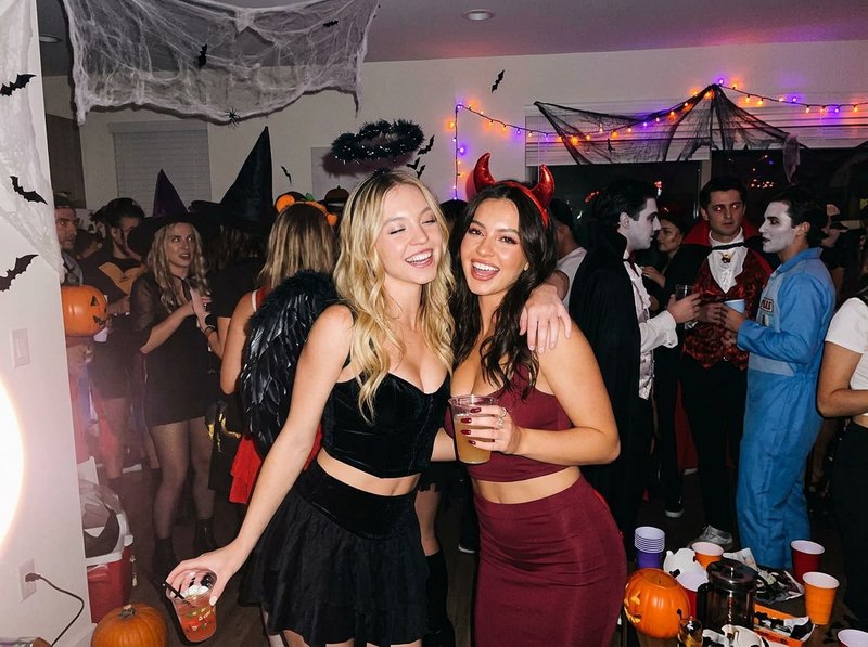 Two smiling friends in Halloween costumes hold drinks while posing at a crowded home party