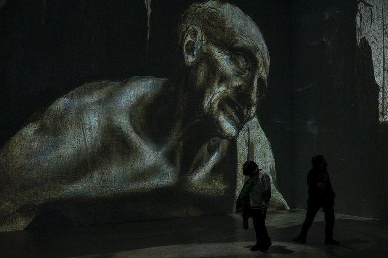Two museum visitors stand beneath a giant projected classical bust of an elderly gaunt man