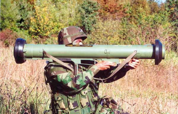 Soldier in camouflage using a green shoulder-fired rocket launcher in a field.