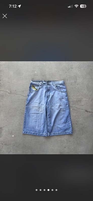 Light wash baggy long denim jean shorts (jorts) laid flat on gray cracked concrete, with a small ...