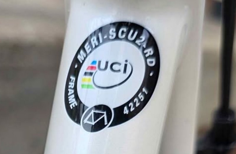 Close-up of a white bicycle frame with a circular UCI label and rainbow stripes.