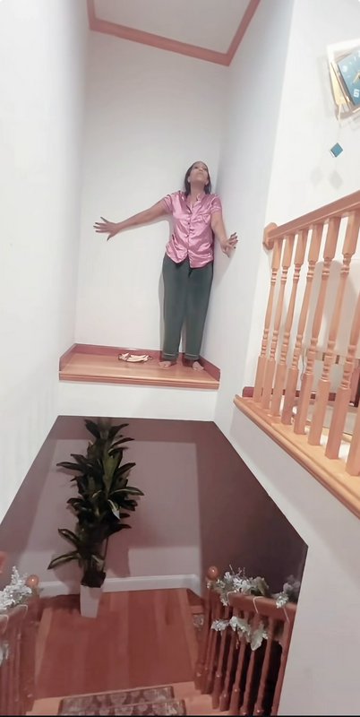 Woman in a pink blouse stands with arms outstretched on a small landing at the top of a wooden staircase.