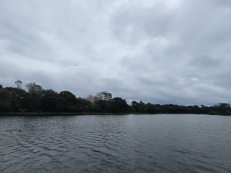 Calm rippled lake under heavy overcast grey clouds, with dense green trees and distant residentia...