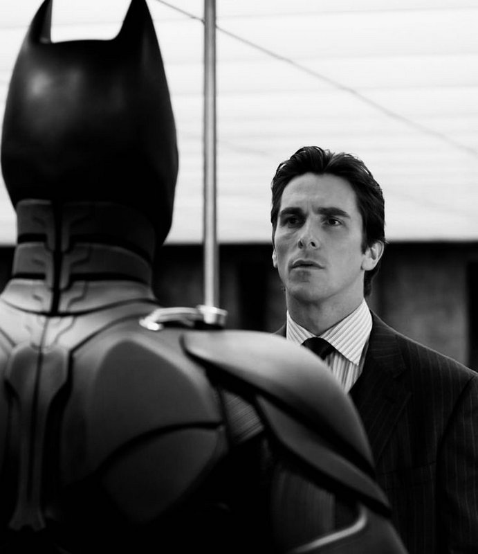 Christian Bale as Bruce Wayne gazes thoughtfully at the back of his armored Batman suit in a moody black and white on-set ...