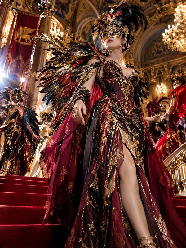 Woman in elaborate feathered masquerade gown walks down red carpeted palace stairs at a lavish ca...