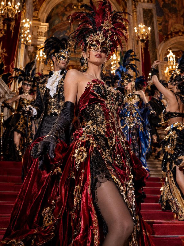 Woman in elaborate red and gold masquerade gown leads masked guests down a grand palace staircase