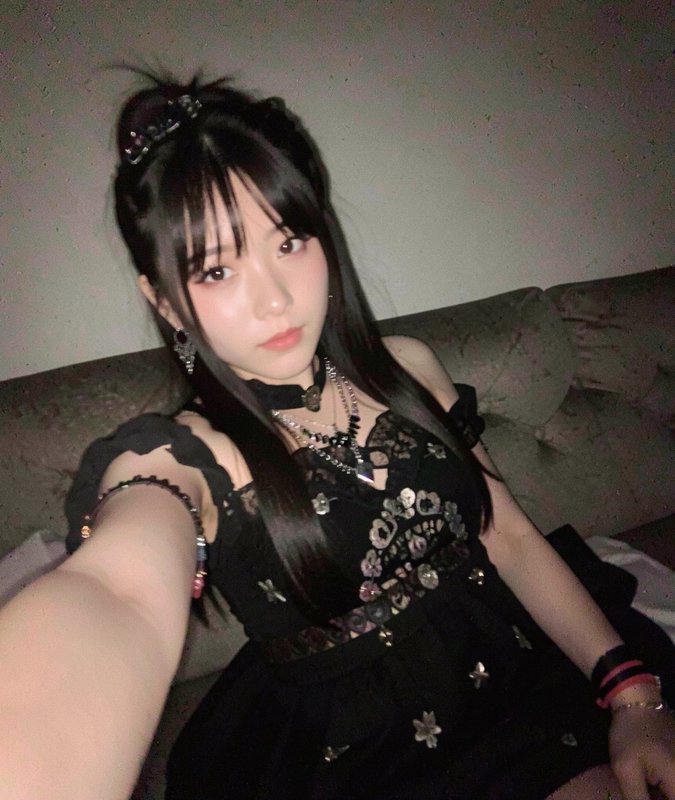 Young woman in a black gothic dress with lace and floral details posing on a couch, taking a selfie.
