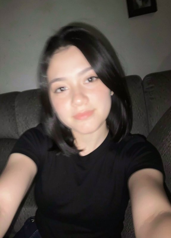 Slightly blurry casual selfie of a young woman with short black bob hair sitting on a dark sofa, ...