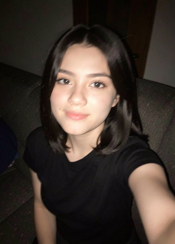 Candid selfie of a young woman with short dark bob hair seated on a dark couch, soft smile in dim...