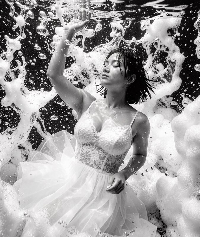 Black and white underwater portrait of a woman in a lace tulle dress reaching upward surrounded b...