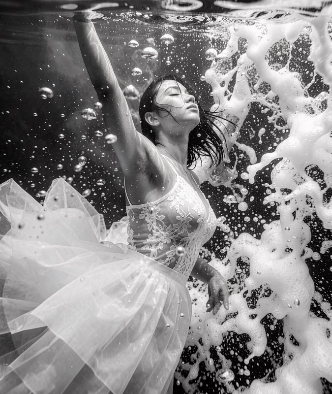 Black and white underwater portrait of a woman in a lace tulle dress floating serenely surrounded by bubbles and foam.