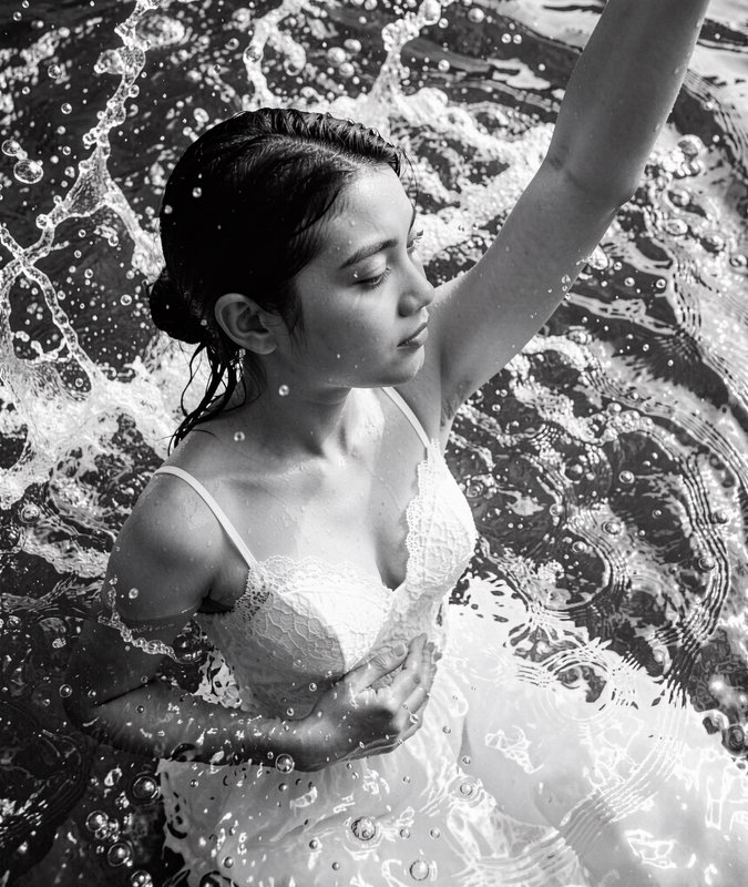 Black and white fine art portrait of a woman in a lace dress standing in splashing rippled water,...