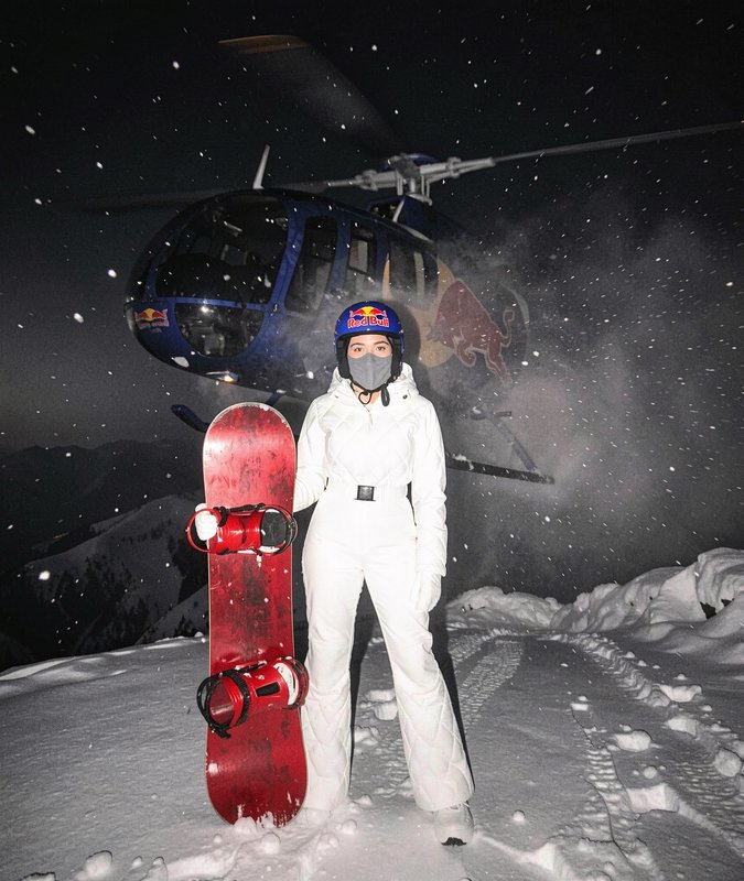 Female pro snowboarder in a white snowsuit holds a red snowboard in front of a Red Bull helicopte...