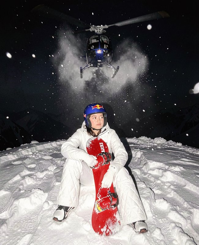 Pro female snowboarder sits on snowy night slope holding a red snowboard, as a hovering helicopte...