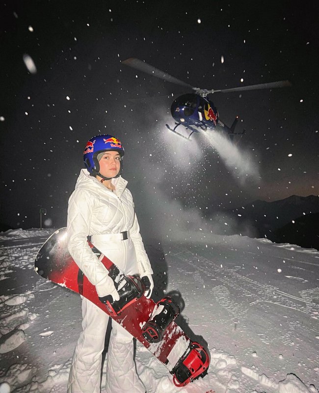 Female Red Bull sponsored snowboarder stands on a snowy night mountain holding her red snowboard,...