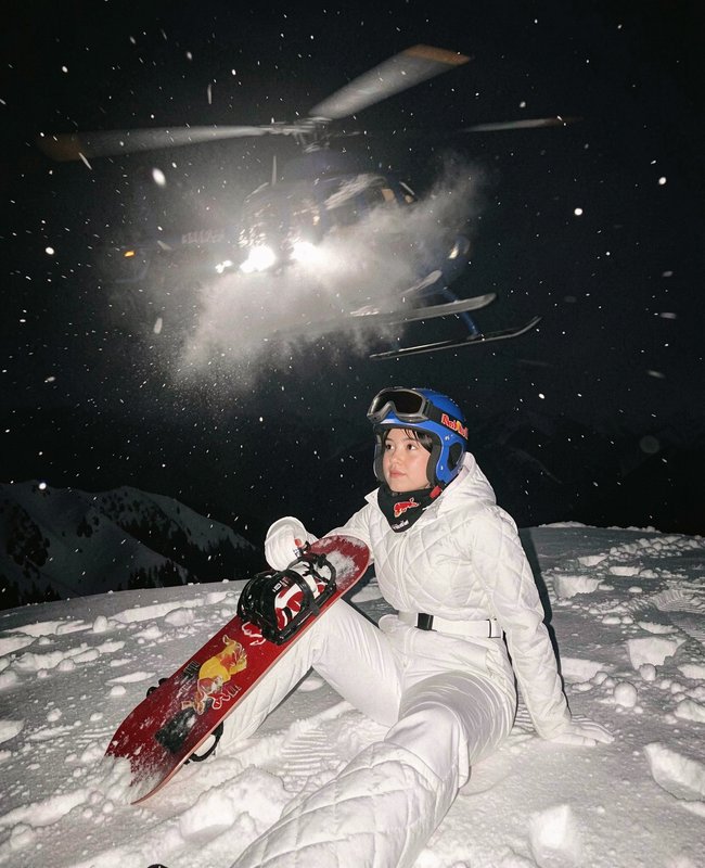 Female heli-snowboarder sits on a snowy mountain peak as a helicopter hovers overhead during nigh...