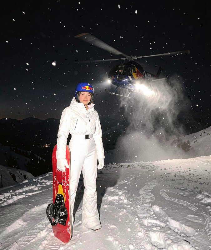 Female snowboarder in a white snowsuit stands on a snowy mountain at night, as a Red Bull helicop...