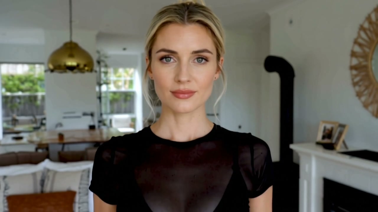 Blonde woman in a sheer black top stands in a bright modern open-concept living room, looking dir...