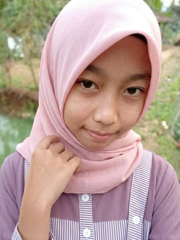 Close-up portrait of a young Muslim girl in a soft pink hijab, posing outdoors near a rural stream