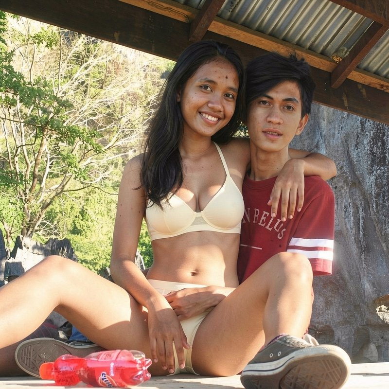 Smiling young Southeast Asian couple sit closely embracing in a sunny outdoor shelter, posing for...